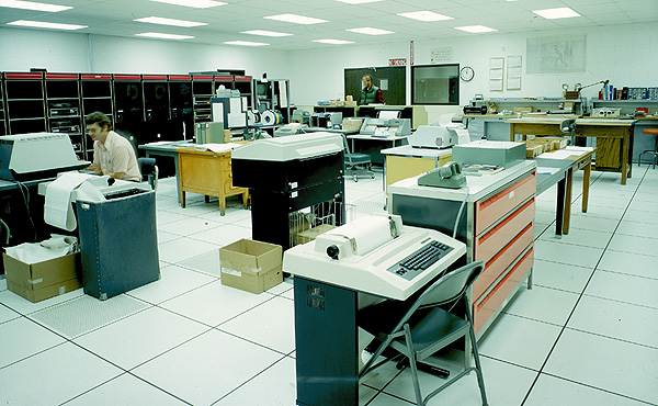 A room a couple of people, several teletype terminals, and a large computer. A teletype terminal consists of a case with a keyboard, printer, and a wide roll of paper. The DEC PDP-11/20 computer equipment takes up almost the entire left wall, it is black with red accents.