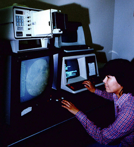 A woman using a text-based computer and showing an image of the moon on a screen next to it.