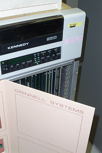 A computer with a dark transparent window on the top corner and the model, KENNEDY 9610. It has a control panel with a switch, 8 buttons, 9 LEDs, and an 8 character display.  It sits on top of a box labeled Grinnel Systems containing vertical electronics racks.